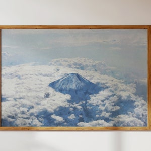 May include: A framed watercolor painting of Mount Fuji, a snow-capped mountain peak, rising above a sea of white clouds. The sky is a soft blue.