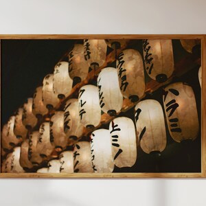 May include: A close-up of a row of white paper lanterns with black Japanese characters hanging from a wooden structure. The lanterns are illuminated from within, casting a warm glow.