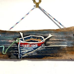 Puede incluir: Adorno de pared de madera flotante pintado a mano con un barco azul y rojo con la inscripción "LOST LOVE" y remos blancos. Una cuerda verde y azul está atada. La madera está envejecida.