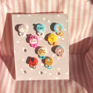 Peut inclure: Une collection d'épingles colorées de style dessin animé représentant divers animaux, dont un hamster, un lapin, un poussin et une coccinelle. Les épingles sont fixées sur une carte grise clair à pois blancs.