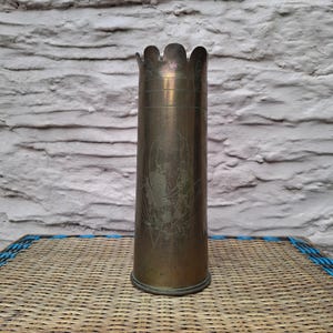 May include: A cylindrical brass artillery shell casing repurposed as a vase, featuring a scalloped top edge and a faint, etched floral design on the front surface. The metal has a weathered, aged patina with visible horizontal lines near the rim.