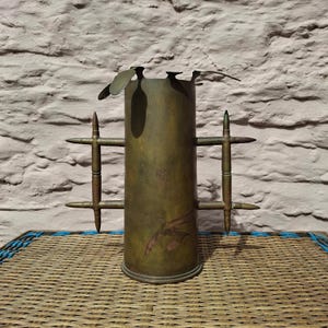 May include: A cylindrical brass vessel featuring two handles constructed from metallic bullet casings. The main body has a weathered, dark patina with a smooth finish. The top rim is adorned with small, flat, leaf-shaped metal accents.