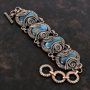 Peut inclure: Bracelet artisanal avec des pierres bleues ovales serties dans un fil de cuivre complexe. Le bracelet est orné de volutes décoratives et d'un fermoir à bascule. Les pierres ont une irisation bleue et verte.