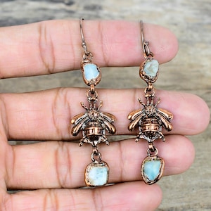 Pode incluir: Um par de brincos cor de cobre com um design de abelha. Cada brinco tem uma pedra azul clara na parte superior e inferior. Os brincos são presos por um gancho.