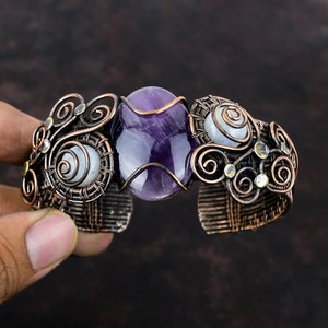以下が含まれることがあります： アメジストの宝石と2つの真珠貝の石が付いた、銅線で巻かれたブレスレット。ブレスレットは、複雑なワイヤーデザインで飾られています。
