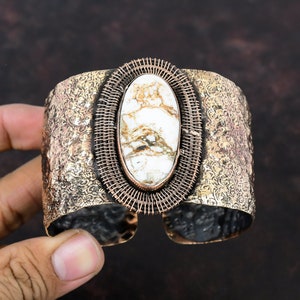May include: A wide copper cuff bracelet with a white and brown oval gemstone set in a wire cage. The bracelet has a hammered texture.
