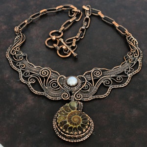 May include: A copper wire necklace with a delicate, intricate design. The necklace features a large, round pendant with a fossilized ammonite and a white pearl in the center.