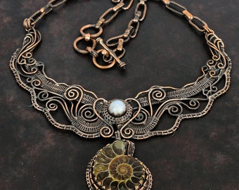 May include: A copper wire necklace with a delicate, intricate design. The necklace features a large, round pendant with a fossilized ammonite and a white pearl in the center.