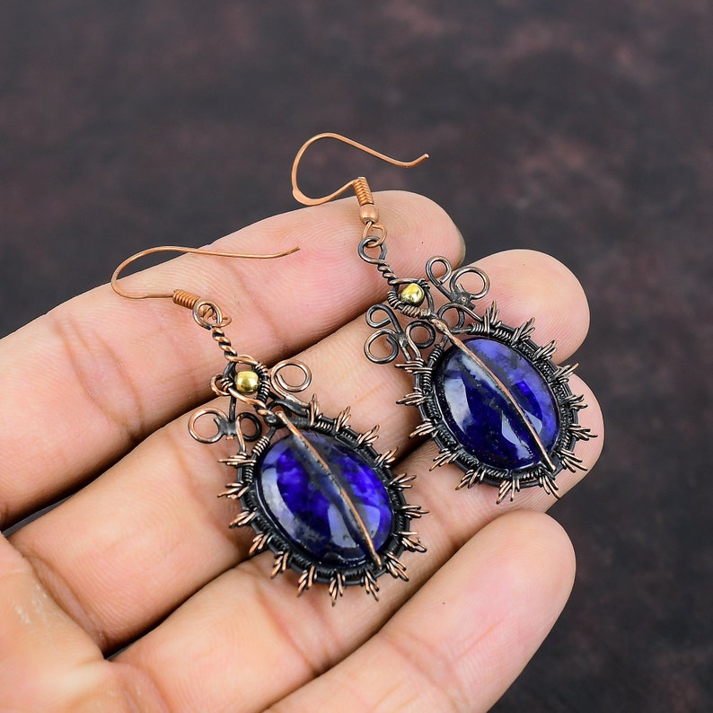 May include: Handmade copper earrings featuring oval, deep blue gemstones. The gemstones are set within a wire frame with a sunburst design. The earrings have a hook closure and small gold-colored accents. The earrings are held in a hand.