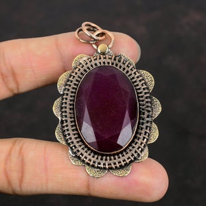 Könnte beinhalten: Ein Anhänger mit einem violetten Edelstein und einem detaillierten, geflochtenen Metallrahmen. Der Anhänger hat einen gewellten Rand und eine kupferfarbene Oberfläche.