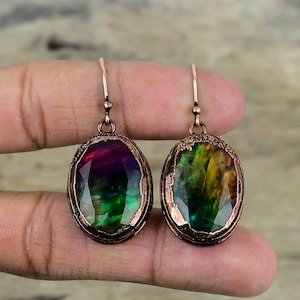 May include: A pair of copper-colored earrings with oval-shaped, iridescent gemstones. The gemstones display a spectrum of colors, including purple, green, and yellow. The earrings have a textured copper frame and a simple hook design.