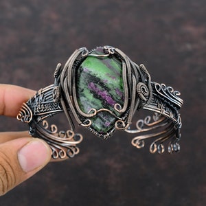 May include: A copper wire wrapped bracelet featuring a green, pink, and black gemstone. The bracelet is crafted with copper wire that is twisted and woven together to create a distinctive design.