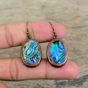 May include: A pair of oval-shaped earrings with a copper frame and iridescent abalone shell inlay. The earrings have a simple hook closure.