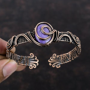 Pode incluir: Um bracelete envolto em fio de cobre com uma gema púrpura e verde. O bracelete é feito com um design de fio tecido e tem um padrão de espiral em torno da gema.