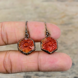 May include: A pair of dangle earrings with a hexagonal red and orange glass flower design. The earrings have a silver hook.