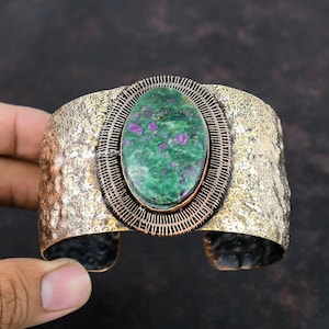 May include: A wide copper cuff bracelet featuring a green and purple oval gemstone set in a wire cage. The bracelet has a hammered texture.