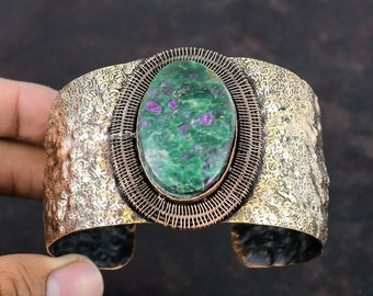 May include: A wide copper cuff bracelet featuring a green and purple oval gemstone set in a wire cage. The bracelet has a hammered texture.