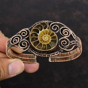 Puede incluir: Una pulsera envuelta en alambre de cobre con un amonite fosilizado en el centro. El amonite es marrón y amarillo con un patrón en espiral. La pulsera tiene un diseño decorativo de espirales.