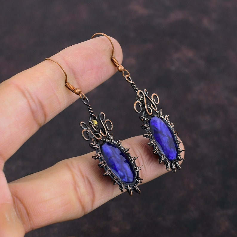 May include: A pair of handcrafted earrings featuring oval, deep blue gemstones. The earrings have a dark copper-colored metal frame with intricate filigree and a spiky border around the gemstones. The ear wires are a lighter copper color.