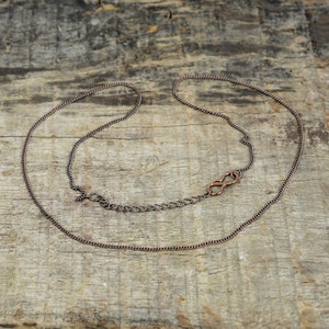 May include: A copper chain necklace with a simple infinity clasp. The chain is lying on a wooden surface.