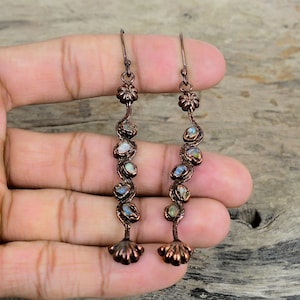 May include: A pair of long, copper earrings with a floral design at the top and a series of iridescent stones hanging down. The stones are set in a wavy, copper wire.