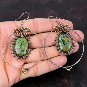 May include: A pair of copper hoop earrings with green and pink gemstone cabochons set in wire wrapped designs. The earrings are approximately 2 inches in diameter.