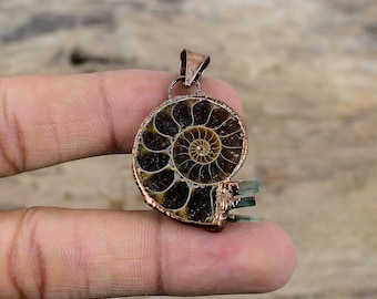 Puede incluir: Un colgante color cobre con una concha de amonita fosilizada con un patrón en espiral. El colgante está adornado con pequeños cristales de turmalina verde claro. La amonita está enmarcada en cobre, creando una pieza de joyería única.
