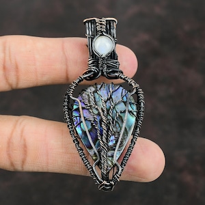 May include: A wire-wrapped pendant featuring a blue and green abalone shell with a tree design. The pendant is accented with a white moonstone.
