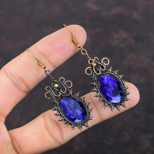May include: A pair of handcrafted earrings featuring large, oval, deep blue gemstones. The stones are set in a dark metal frame with a sunburst design. The earrings have intricate wirework and copper-colored ear wires.