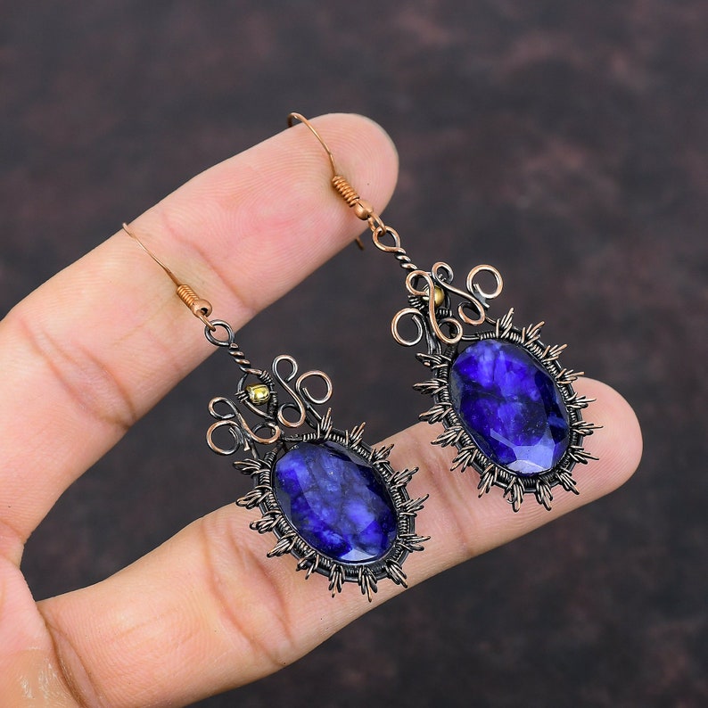 May include: A pair of handcrafted earrings featuring large, oval, deep blue gemstones. The stones are set in a dark metal frame with a sunburst design. The earrings have intricate wirework and copper-colored ear wires.