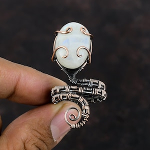May include: A copper wire wrapped ring with a white moonstone cabochon. The ring is designed in a spiral shape with a delicate wire design.