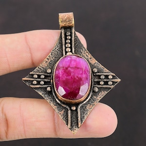 May include: A pendant featuring a large, oval-cut ruby gemstone set in a diamond-shaped, ornate bronze frame. The frame has a textured surface with decorative patterns and a loop for a chain. The ruby is a vibrant shade of pink.