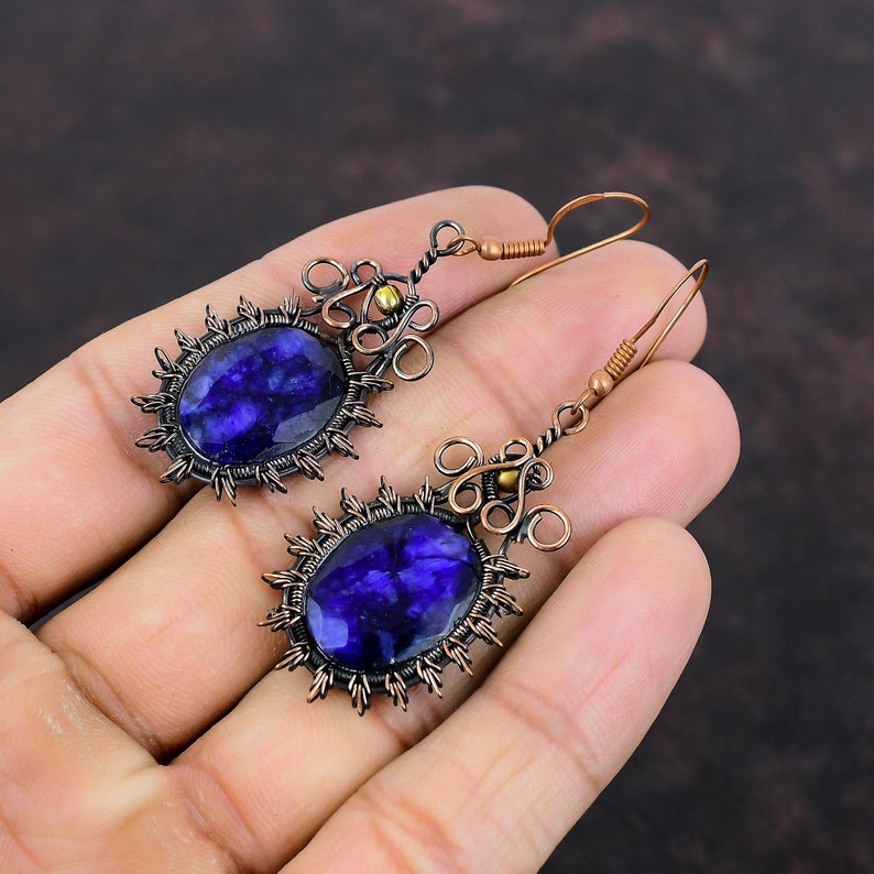 May include: A pair of handcrafted earrings featuring large, oval, deep blue gemstones. The gemstones are set in a dark metal frame with a sunburst design. The earrings have copper-colored wire detailing and ear wires, creating a unique, artisan look.
