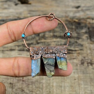Puede incluir: Aretes de aro de cobre con una barra de cobre martillada y piedras de labradorita colgantes. La barra tiene un pequeño diseño de cruz y el aro está acentuado con piedras turquesa.