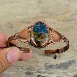May include: Copper bracelet with a heart-shaped design and a large oval-shaped stone with a mix of blue, green, and brown crystals.
