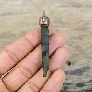 Puede incluir: Un colgante largo de piedra verde oscuro y negro con una parte superior envuelta en alambre de cobre y un pequeño acento de turquesa.