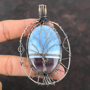 May include: A copper wire wrapped pendant featuring a blue and purple agate stone with a tree design. The pendant is oval shaped and has a copper bail.