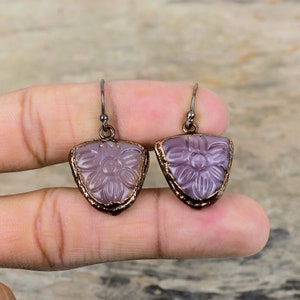 Könnte beinhalten: Ein Paar Ohrringe mit einem violetten Blumendesign. Die Ohrringe sind aus Metall und haben eine kupferne Oberfläche. Die Blumen sind in das Metall geschnitzt und haben ein zartes, feminines Aussehen.