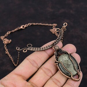 K&ouml;nnte beinhalten: Handgefertigte Kupferkette mit einem drahtumwickelten, ovalen Pyritstein-Anh&auml;nger. Die Halskette hat eine filigrane Kette und ein einzigartiges, k&uuml;nstlerisches Design. Der Pyritstein hat ein metallisches, gesprenkeltes Aussehen.