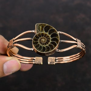 May include: Copper cuff bracelet featuring a fossilized ammonite. The bracelet has a three-strand design with decorative accents on the sides. The ammonite is a spiral shape with shades of brown and green.