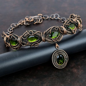 Op de afbeelding: Een ketting van gewikkeld koperdraad met een hanger met groene edelstenen. De ketting heeft een delicate ketting en is versierd met ingewikkeld draadwerk.