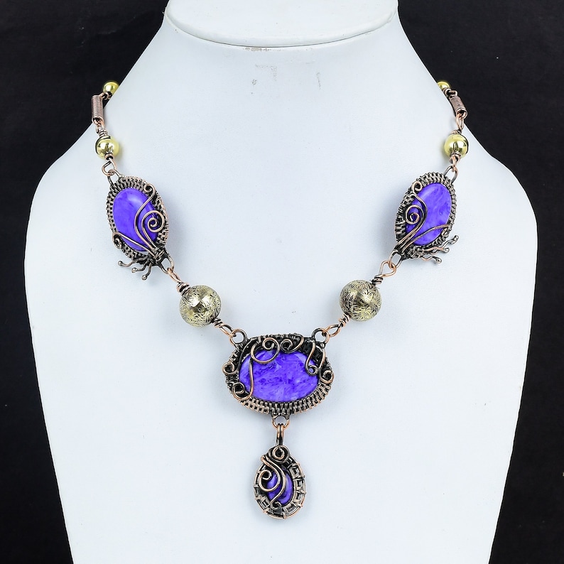 May include: A handcrafted necklace featuring a series of oval and teardrop-shaped purple stones. The stones are set in ornate, dark copper-colored wirework with decorative accents. The necklace is displayed on a white mannequin.