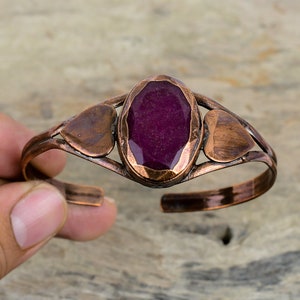 May include: A copper bracelet with a large, oval-shaped, dark purple gemstone set in the center. The bracelet is accented with two smaller heart-shaped copper pieces on either side of the gemstone.