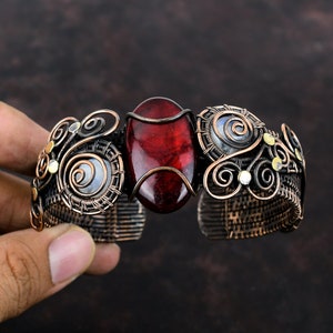 Op de afbeelding: Een armband van koperdraad met een rode edelsteen en witte iriserende stenen. De armband is gemaakt met ingewikkeld draadwerk en heeft een delicaat, vrouwelijk ontwerp.