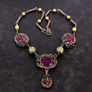 May include: A copper wire wrapped necklace with three purple gemstones and gold beads. The necklace has a pendant with a purple gemstone.