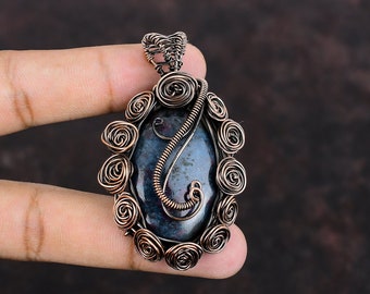 May include: A copper wire wrapped pendant featuring a dark blue and black gemstone with copper wire accents. The pendant is oval shaped and has a wire bail for hanging.