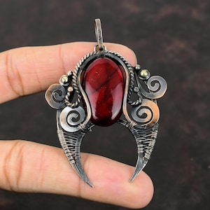 May include: A red gemstone pendant with a copper wire design in the shape of a crescent moon. The pendant has a detailed wire design with swirls and a small gold bead accent.