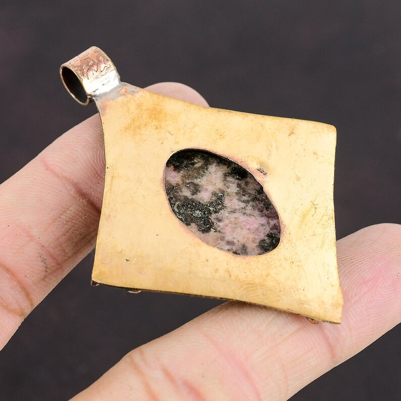 May include: A brass pendant with a diamond shape and a round opening in the centre. The opening is filled with a pink and black stone.