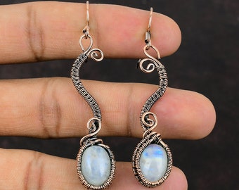Puede incluir: Un par de pendientes de alambre de cobre con una piedra ovalada de piedra lunar en cada uno. Los pendientes son largos y colgantes, con un diseño delicado de alambre.