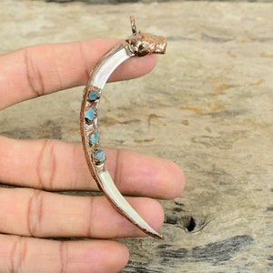 Może przedstawiać: Wisiorek w kształcie półksiężyca wykonany z białej muszli z miedzianymi akcentami i niebieskimi kamieniami szlachetnymi. Wisiorek jest przymocowany do miedzianej zawieszki.